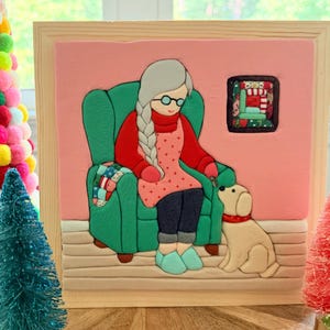 May include: A framed clay art piece featuring a person with long gray hair in a braid, wearing glasses, a red sweater, and a pink patterned apron, sitting in a green armchair. A small cream-colored dog sits beside the chair on a beige floor.