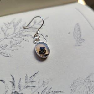 May include: A silver earring with a small, oval-shaped pendant. The pendant features a clear, light-colored stone with a black and brown dendritic inclusion, resembling a miniature tree. The earring hangs from a simple hook.