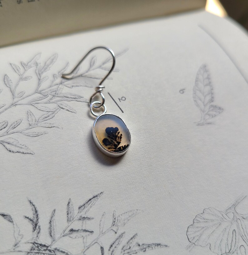May include: A silver earring with a small, oval-shaped pendant. The pendant features a clear, light-colored stone with a black and brown dendritic inclusion, resembling a miniature tree. The earring hangs from a simple hook.