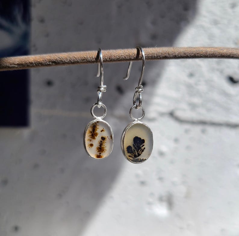 May include: A pair of oval-shaped earrings with silver-colored hooks. Each earring features a clear, light-colored stone with unique inclusions. One earring has brown, tree-like patterns, while the other has a dark blue floral design. The earrings are displayed on a brown stick.