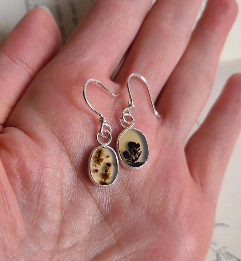 May include: A pair of silver dangle earrings. Each earring features an oval-shaped, bezel-set stone with a unique, natural pattern in shades of brown and cream. The earrings have a simple, elegant design.