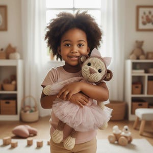 Op de afbeelding: Een kind houdt een handgemaakte gehaakte apenpop vast. De pop heeft een beige gezicht, bruine oren en een roze tutu. Het kind draagt een roze shirt en glimlacht. De achtergrond omvat een raam en een witte plank.