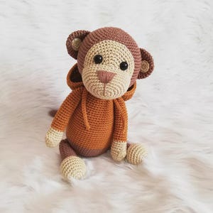 May include: A handmade crochet monkey doll in shades of brown, tan, and orange. The monkey has a hood, large black eyes, and a tan face and hands. The doll is sitting on a white, fluffy surface.
