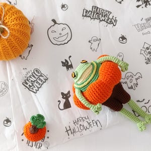 Könnte beinhalten: Ein handgefertigtes Halloween-Häkelset mit einem orangefarbenen Kürbis, einem froschartigen Wesen und einem kleinen Kürbis. Die Artikel liegen auf einer weißen Oberfläche mit Halloween-Zeichnungen und dem Text "HAPPY HALLOWEEN".
