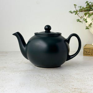 May include: A matte black ceramic teapot with a rounded body, a curved spout, a matching lid with a small knob handle, and a C-shaped handle. The teapot features a smooth, solid dark finish and a classic, symmetrical design for brewing tea.