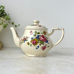 May include: A cream-colored ceramic teapot featuring a ribbed body and a floral pattern with pink, yellow, and blue flowers. The teapot includes a matching lid with a gold-trimmed handle, spout, and rim, showcasing a classic design.