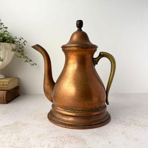 May include: A copper teapot with a curved spout, a brass handle, and a dark brown knob on the lid. The teapot has a wide, rounded body that tapers toward the top and sits on a circular base. The metal surface shows a warm, metallic finish.