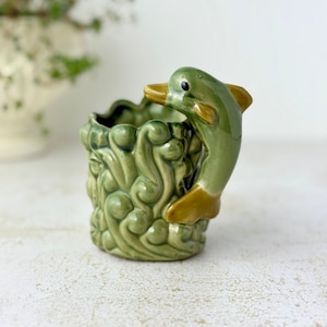 May include: A ceramic mug featuring a glossy green finish with a sculpted dolphin handle. The body of the mug is decorated with a raised, swirling wave pattern in shades of light and dark green, while the dolphin handle has brown accents.
