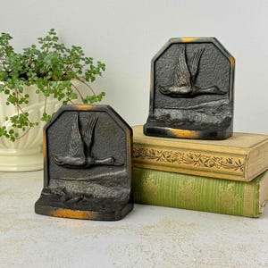 May include: A pair of dark, heavy metal bookends featuring a raised relief design of a flying duck in flight. The bookends have a textured, matte black finish with subtle gold-toned highlights along the edges and the duck's wings.