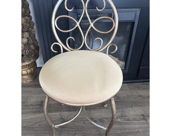 Silla de tocador vintage de metal con respaldo ornamentado en forma de voluta y asiento acolchado color oro champán.