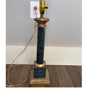 Puede incluir: Una lámpara de mesa decorativa con una columna de mármol verde oscuro y una base cuadrada. La lámpara presenta detalles dorados en la base, la columna y la parte superior. La lámpara está enchufada a una toma de corriente y tiene un cable marrón.