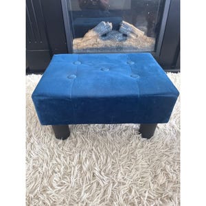 May include: A square, tufted ottoman in a deep blue velvet fabric. The ottoman has six button details and sits on four short, black cylindrical legs. The piece is placed on a white, shaggy rug.