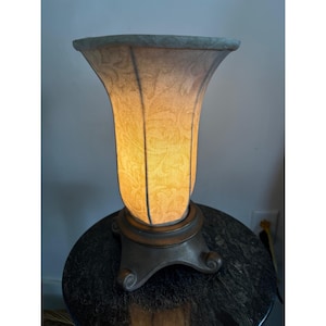 Op de afbeelding: Een decoratieve tafellamp met een patroon lampenkap. De lampenkap is lichtgeel van kleur met een bloemenmotief. De basis is donkerbrons van kleur met een decoratief, gebogen voetontwerp.