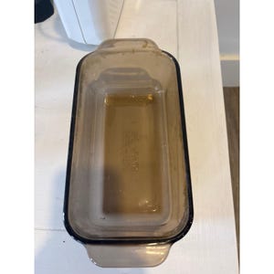 Peut inclure: Un moule à pain rectangulaire en verre brun, avec des coins arrondis et deux poignées. Le moule est vide et semble propre. Il est posé sur une surface blanche.