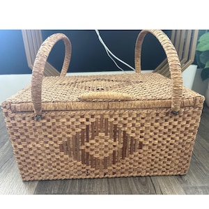 May include: A rectangular woven picnic basket with a lid and two arched handles. The basket is made of light brown woven material with a diamond pattern on the front. The basket is sitting on a wooden surface.