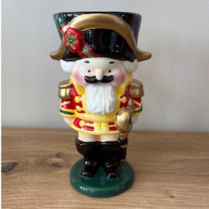 May include: A ceramic nutcracker figurine in the shape of a soldier. The figurine has a black hat with gold trim, a red and gold jacket, and black boots. The base is green. The nutcracker is a decorative item.