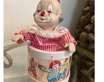 Può includere: Un giocattolo vintage a carica con un clown che esce da un tamburo. Il clown ha una faccia dipinta, un naso rosso e una camicia a righe rosse e bianche. Il tamburo è decorato con illustrazioni di clown e ha una combinazione di colori bianco e rosa.