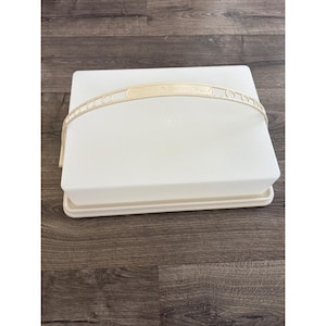 Könnte beinhalten: Ein rechteckiger, weißer Tupperware-Kuchenbehälter mit flachem Boden und einem hohen, abnehmbaren Deckel. Der Behälter verfügt über einen cremefarbenen, gewölbten Griff, der über die Oberseite verläuft und für den Transport von Backwaren dient.
