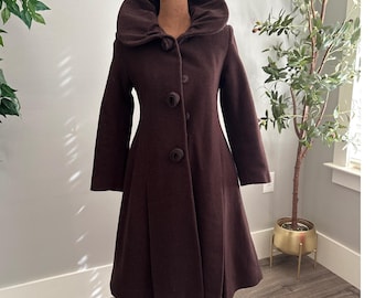 Puede incluir: Un abrigo marrón oscuro de estilo vintage con un gran cuello redondo y tres botones decorativos. El abrigo tiene una falda acampanada y mangas largas. El abrigo se exhibe en un maniquí.