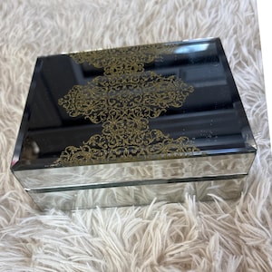 以下が含まれることがあります： 黒い天板と金色の透かし模様が施された、鏡張りのジュエリーボックス。 ボックスは長方形で、ふわふわの白い表面に置かれています。 金色の透かし模様は、装飾的で繰り返しのパターンです。