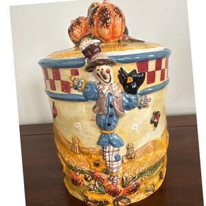 Op de afbeelding: Een keramische koekjespot met een herfstthema. De pot heeft een lachende vogelverschrikker in een blauwe outfit, omringd door pompoenen, zonnebloemen en herfstbladeren. De deksel is bekroond met twee oranje pompoenen.