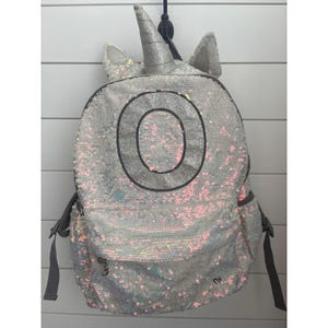 May include: A silver sequined backpack with a unicorn horn and ears. The backpack has a large letter "O" on the front and a small heart detail. Gray straps and a front pocket complete the design.