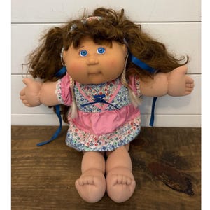 May include: A vintage Cabbage Patch Kid doll with brown hair, blue eyes, and rosy cheeks. The doll wears a pink dress with a floral print bodice and white lace trim. Blue ribbons are tied in the hair and on the dress.