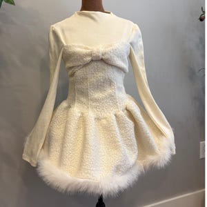 Puede incluir: Vestido color marfil con mangas largas y cuello alto. El corpiño presenta un lazo texturizado y una cintura entallada, que desemboca en una falda acampanada adornada con piel sintética blanca. El vestido se exhibe en un maniquí.