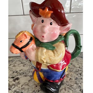 Puede incluir: Una tetera de cerámica caprichosa con forma de elfo jovial sosteniendo un caballo de juguete. El elfo lleva un sombrero rojo, una bufanda verde y ropa colorida. La tetera tiene un asa y un pico verdes.