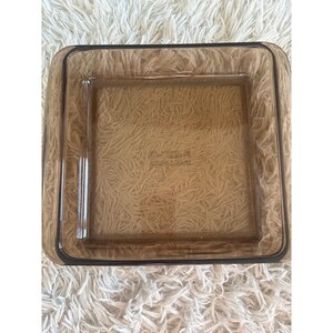 Puede incluir: Un plato cuadrado de vidrio marrón con un borde ligeramente redondeado. El plato tiene forma rectangular con un interior cuadrado. El vidrio es transparente, lo que permite ver la superficie debajo. El plato probablemente se utiliza para servir o exhibir.