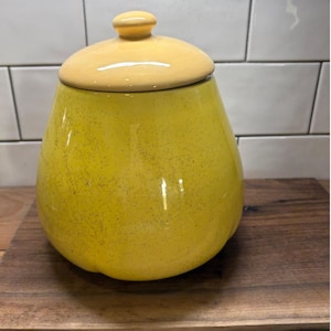 Puede incluir: Un tarro de cerámica amarillo con tapa a juego. El tarro tiene forma redondeada y textura moteada. La tapa es de un tono amarillo más claro y tiene un pequeño pomo en la parte superior. El tarro está sobre una superficie de madera.