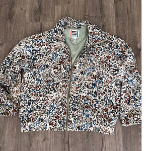 Puede incluir: Chaqueta con cremallera y estampado colorido. La chaqueta presenta un patrón de manchas marrones, azules y blancas sobre un fondo crema. Tiene un cuello alto y cierre de cremallera. La chaqueta está colocada sobre una superficie de madera.