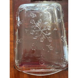 Puede incluir: Fuente rectangular de vidrio transparente con diseño floral en relieve. La fuente presenta un motivo floral central y flores más pequeñas en los bordes. El vidrio transparente permite ver el contenido.
