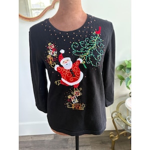 May include: Black Christmas sweater with a festive Santa Claus design. The sweater features an embroidered Santa Claus, a green Christmas tree, and gift boxes. The neckline is embellished with small, gold-colored sequins. A red bird is perched on the Christmas tree.