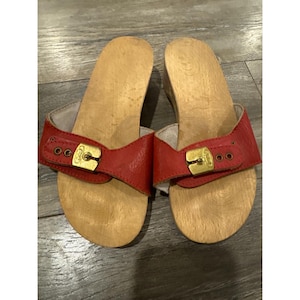 Könnte beinhalten: Ein Paar Holzclogs mit roten Lederriemen. Die Riemen haben goldfarbene Schnallen und sind mit Nieten befestigt. Die Clogs haben eine natürliche Holzoberfläche und einen leichten Absatz. Diese Clogs sind ein klassischer Schuhstil.