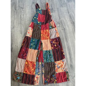 以下が含まれることがあります： さまざまな色の柄の生地の正方形を組み合わせたパッチワークジャンプスーツ。ジャンプスーツはワイドレッグ、調節可能なストラップ、フロントポケット付き。生地の正方形には、赤、オレンジ、緑、茶色の花柄、ストライプ、ペイズリー柄が含まれています。