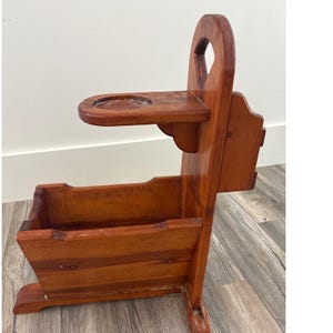 Può includere: Un portapipe vintage in legno con una calda finitura marrone-rossastra. È dotato di un posacenere integrato, un vano portaoggetti e un manico. Il supporto è progettato per contenere una pipa e del tabacco, mostrando un'estetica rustica classica.