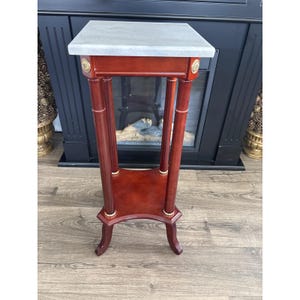 May include: A tall, square pedestal stand featuring a white marble top and a polished reddish-brown wooden frame. The structure includes four slender legs with gold-toned circular accents at the top and base, connected by a lower square shelf.