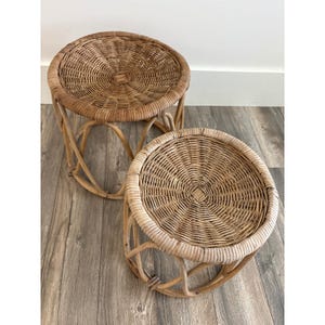 May include: Two round, woven rattan side tables. The tables have a natural, light brown color with a woven top and curved legs. The tables are placed on a wooden floor.