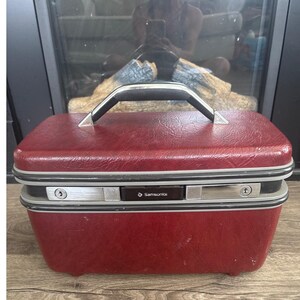 May include: A vintage red Samsonite train case with a black handle and silver accents. The rectangular case has a textured surface, a silver frame, and two silver latches. The Samsonite logo is visible on the front.