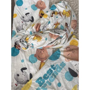 May include: White bedsheets featuring cartoon characters Snoopy and Charlie Brown, with blue polka dots. The fitted sheet and pillowcase are visible, with the words "Best Friends" printed on the pillowcase. The sheets are made of a soft, lightweight fabric.