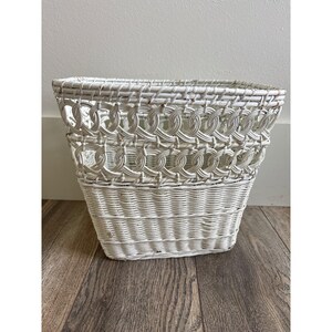 May include: A white wicker basket with a rectangular shape. The basket features a woven design with a decorative chain-link pattern around the middle. The basket is suitable for storage and home decor.