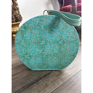 Könnte beinhalten: Eine runde Vintage-Handtasche mit einem türkis-grünen Blumenmuster. Die Tasche hat einen passenden türkisfarbenen Griff und einen silberfarbenen Metallverschluss. Die Tasche steht auf einem Holzboden.