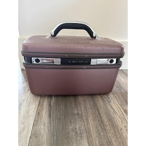 May include: A vintage Samsonite train case in a deep burgundy color. The case has a rectangular shape with a curved handle and silver-toned hardware. The case is sitting on a wooden floor.