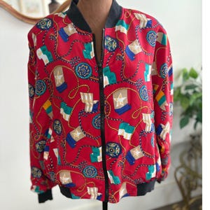 May include: A red bomber jacket with a navy blue collar and cuffs. The jacket features a repeating pattern of flags, stars, and nautical rope designs in white, blue, green, and gold. The jacket has a black zipper closure.