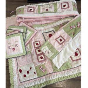 May include: A quilted bedding set featuring a patchwork design in sage green, soft pink, and white. The fabric displays embroidered ladybugs, dragonflies, and flower motifs. The set includes a long crib bumper, a quilt, and several square pillows.