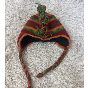 May include: A colorful knit hat with ear flaps and a braided tie. The hat features horizontal stripes in red, orange, and green. A green tassel adorns the top. The hat's interior is black.