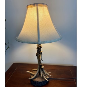 May include: A rustic table lamp with a beige lampshade and a base designed to resemble deer antlers. The lamp has a warm glow, casting soft light. The base is brown and beige, with the antlers extending upwards to support the lampshade.