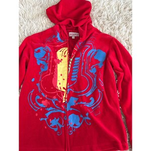 Puede incluir: Una sudadera roja con cremallera y capucha, con un diseño gráfico colorido. El diseño incluye elementos azules, amarillos y rojos. La sudadera tiene una etiqueta que dice "MIRIKA".