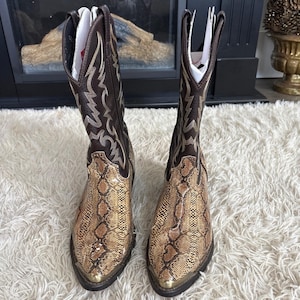 Puede incluir: Un par de botas de vaquero marrones y beige con un estampado de piel de serpiente en el pie y un diseño decorativo cosido en la caña. Las botas tienen una puntera puntiaguda y una punta de metal dorado.